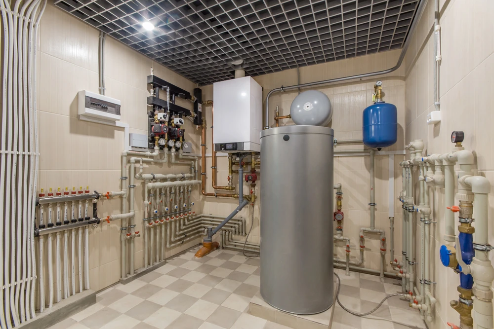 Local technique moderne avec chaudière murale blanche, ballon d'eau chaude cylindrique gris, manifold de distribution avec vannes rouges, tuyauterie en cuivre et PVC blanc, vase d'expansion bleu suspendu