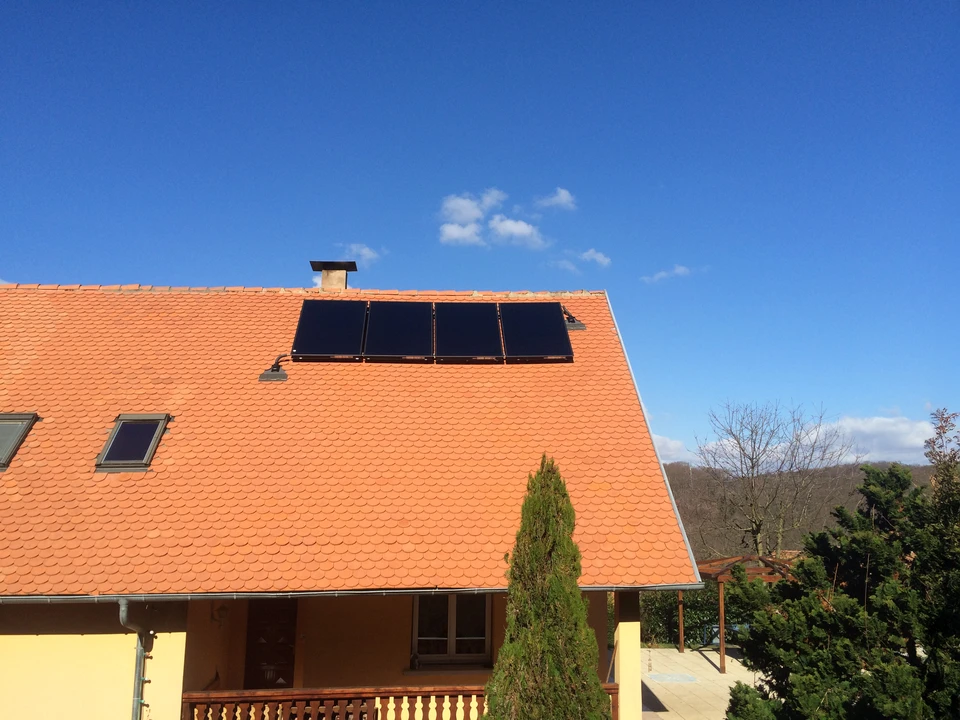 Technicien en combinaison de travail grise et jaune effectuant la maintenance d'une installation solaire photovoltaïque sur toiture, sous ciel bleu nuageux